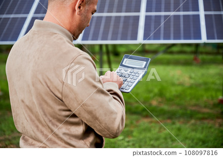 Professional investor calculating his possible profit from alternative enegry sources. Man using calculator to count exact sum of income. Back view of man counting on background of solar panels. Professional investor calculating his possible profit from alternative enegry sources. Man using calculator to count exact sum of income. Back view of man counting on background of solar panels. 108097816