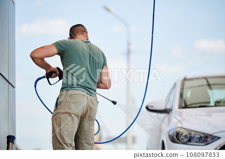 Washing luxury white auto with water gun on an open air car wash. Confident man cleaning his car with high pressure water jet. Technical service concept. 108097833