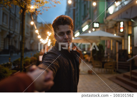 follow me. Beautiful romantic curly man takes his girlfriend hand while walking in the evening city 108101562