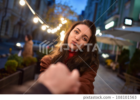 follow me. Beautiful romantic brunette girl in red coat takes her boyfriend's hands while walking in the evening city 108101563