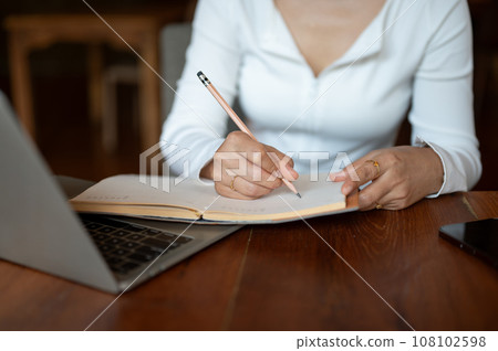 Cropped image of a woman writing some ideas down in her book, working remotely at a coffee shop. 108102598