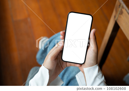 Top view of a woman using her smartphone while sitting indoors. A white-screen smartphone mockup. 108102608