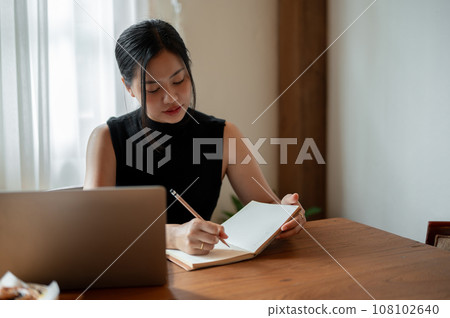A beautiful Asian woman working remotely at a coffee shop is writing some ideas down in her book. 108102640
