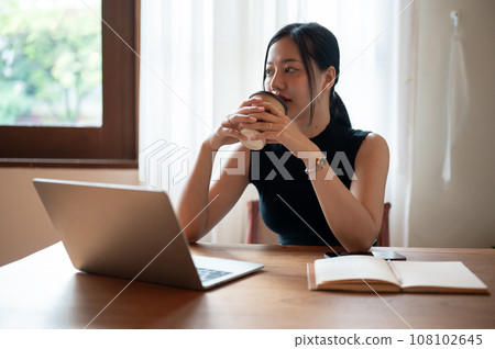 A thoughtful Asian businesswoman is sipping coffee while sitting at a table in a coffee shop. 108102645