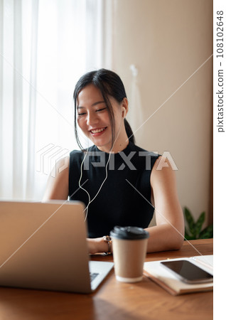 A beautiful Asian woman is having an online meeting while working remotely at a coffee shop. 108102648