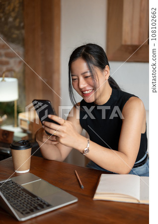 A cheerful Asian woman enjoys watching funny videos online on her phone while sitting in a cafe. A cheerful Asian woman enjoys watching funny videos online on her phone while sitting in a cafe. 108102662