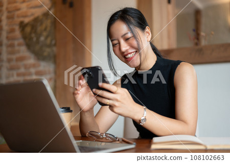 A woman is laughing and enjoys chatting with her friends on her phone while sitting in a cafe. A woman is laughing and enjoys chatting with her friends on her phone while sitting in a cafe. 108102663