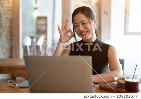 A businesswoman is joining an online meeting, showing the Okay hand sign, working at a cafe. 108102677