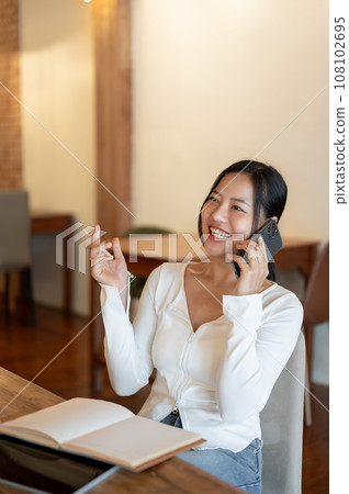 A cheerful Asian woman enjoys talking on the phone with her friend while relaxing in a coffee shop. A cheerful Asian woman enjoys talking on the phone with her friend while relaxing in a coffee shop. 108102695