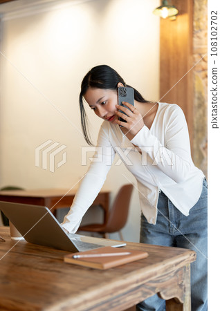 A focused businesswoman is talking on her phone while leaning and working on her laptop on the table 108102702