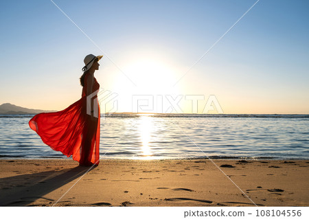 Young woman wearing long red dress and straw hat standing on sand beach at sea shore enjoying view of rising sun in early summer morning. Young woman wearing long red dress and straw hat standing on sand beach at sea shore enjoying view of rising sun in early summer morning. 108104556