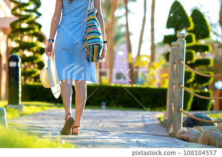 Young woman wearing light blue summer dress holding fashionable shoulder bag and yellow straw hat standing outside enjoying warm weather in summer park. 108104557