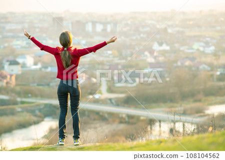 Young woman standing outdoors raising her hands enjoying city view. Relaxing, freedom and wellness concept. Young woman standing outdoors raising her hands enjoying city view. Relaxing, freedom and wellness concept. 108104562