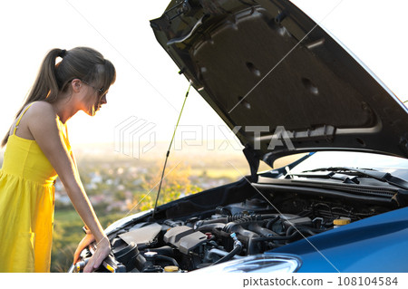 Young woman driver standing near her car with popped hood having engine problems. 108104584