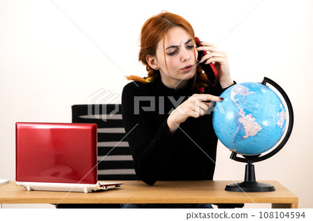 Young travel agent woman sitting behind working desk with laptop computer and geographic globe of the world talking on a cell phone. 108104594