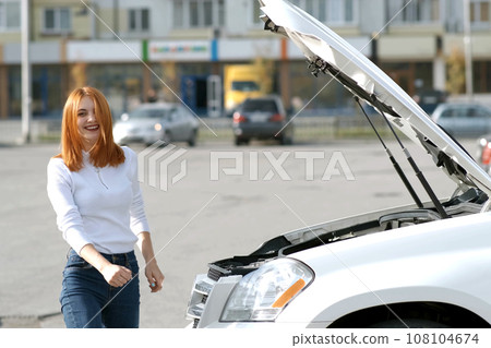 Young funny smiling woman driver near broken car with popped hood having a prbreakdown problem with her vehicle waiting for assistance. 108104674