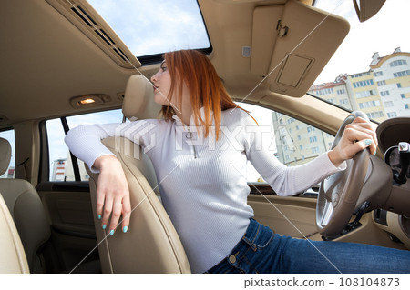Wide angle view of young redhead woman driver driving a car backwards looking behind. 108104873