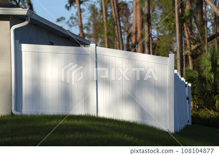White vinyl picket fence on green lawn surrounding property grounds for backyard protection and privacy White vinyl picket fence on green lawn surrounding property grounds for backyard protection and privacy 108104877
