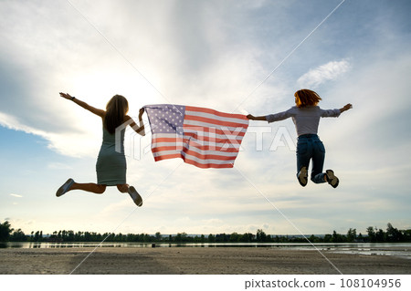 Two young friends women holding USA national flag jumping up together outdoors at sunset. Patriotic girls celebrating United States independence day. 108104956