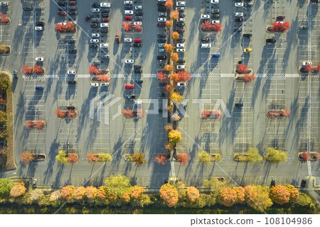 Top view of many cars parked on a parking lot in front of a strip mall plaza. Concept of consumerism and market economy Top view of many cars parked on a parking lot in front of a strip mall plaza. Concept of consumerism and market economy 108104986