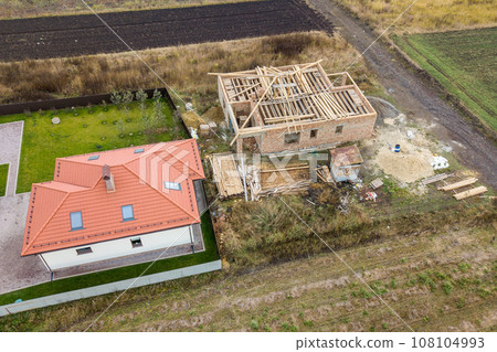 Top down aerial view of two private houses, one under construction with wooden roofing frame and another finished with red tiled roof. 108104993
