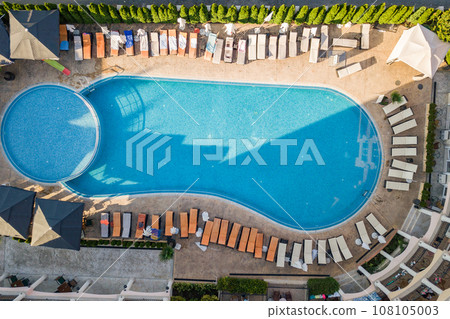 Top down aerial view of hotel swimming pool with crystal blue water surrounded with palm trees and deck laying chairs in resort city near the sea in warm summer season. 108105003