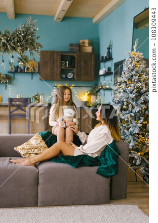 Mother and daughter are sitting on a cozy sofa and playing near the Christmas tree 108109443