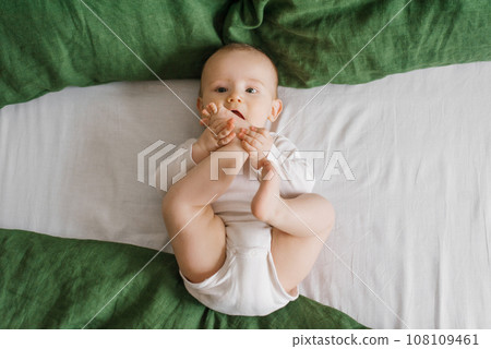 Cute baby is lying on a green and white bedspread on the bed. A smiling child having his feet in a sunny bedroom. 108109461