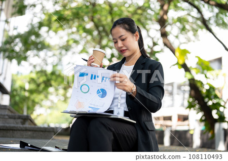 A busy businesswoman is sipping coffee and reviewing business documents while sitting on the stairs. 108110493