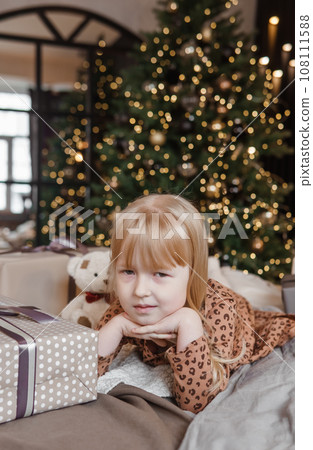 A little blonde girl is lying on a soft bed in a festive interior decorated in a New Year's style. The child is waiting for a New Year's gift. A little blonde girl is lying on a soft bed in a festive interior decorated in a New Year's style. The child is waiting for a New Year's gift. 108111588