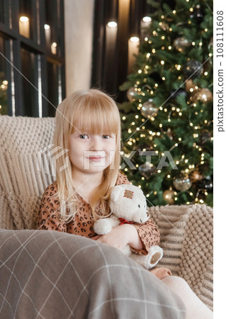 A little blonde girl is sitting in an armchair in an interior decorated in a New Year's style. A child holds a teddy bear in his hands. A little blonde girl is sitting in an armchair in an interior decorated in a New Year's style. A child holds a teddy bear in his hands. 108111608