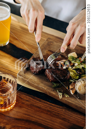 Close up of hands holding knife and fork is cutting grilled ribeye steak on wooden cutting board 108112653
