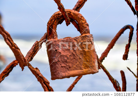 Old ferruginous padlock hanging on a rusty fence on a sea background Old ferruginous padlock hanging on a rusty fence on a sea background 108112877