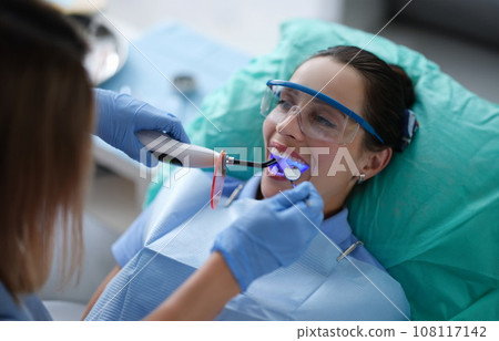 Young woman dentist in protective gloves checking dental seal, fixing photopolymer lamp during stomatology procedure 108117142