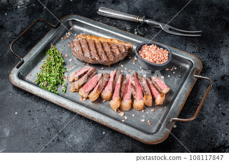 Grilled and sliced brazilian Picanha steak, cup rump beef meat steak on a steel serving tray with herbs. Black background. Top view. Copy space Grilled and sliced brazilian Picanha steak, cup rump beef meat steak on a steel serving tray with herbs. Black background. Top view. Copy space 108117847