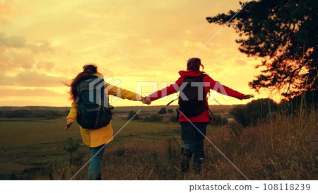 Happy women tourists run and jump joining hands at hike in sunset valley Happy women tourists run and jump joining hands at hike in sunset valley 108118239