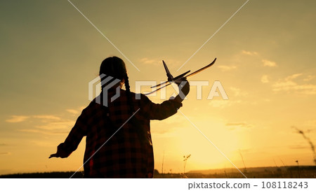 Schoolgirl holds toy plane running in countryside meadow at sunset time 108118243