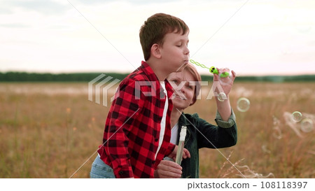 Cheerful boy in checkered shirt blows bubbles while mother helps on mowed field Cheerful boy in checkered shirt blows bubbles while mother helps on mowed field 108118397