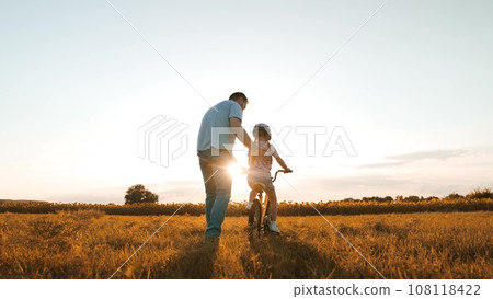 Caring father helps little daughter to ride bicycle in evening meadow on weekend 108118422