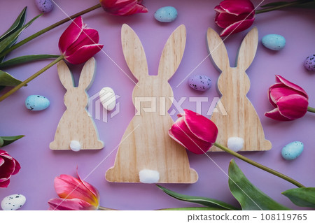 Easter eggs and wooden bunnies with pink tulips on violet pastel background. Flat lay composition, top view. Decoration minimalist modern design template. Floral layout Concept of creative spring Easter eggs and wooden bunnies with pink tulips on violet pastel background. Flat lay composition, top view. Decoration minimalist modern design template. Floral layout Concept of creative spring 108119765