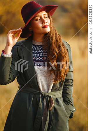 Beautiful stylish girl walks in the autumn park. The girl is dressed in a green coat and a red hat. 108120382