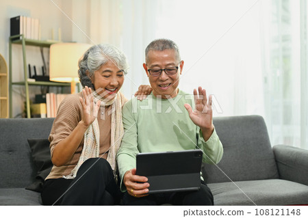 Cheerful elderly man waving hand speaking on video call talking to family while resting on armchair at home 108121148