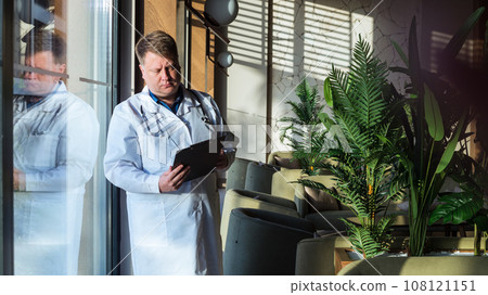 A male doctor with documents in his hands stands near the window looking into the distance A male doctor with documents in his hands stands near the window looking into the distance 108121151