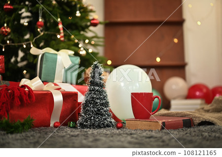 A cup of hot chocolate, pile of gift boxes, coffee cup and book on carpet near decorated Christmas tree A cup of hot chocolate, pile of gift boxes, coffee cup and book on carpet near decorated Christmas tree 108121165