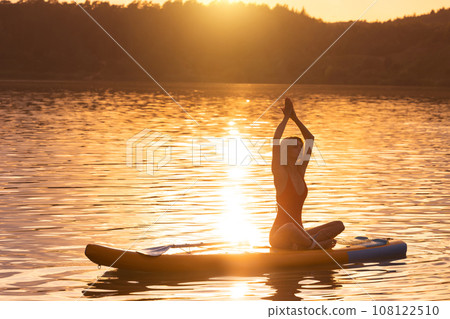 Young woman in yoga pose practicing on paddle sup surfboard during sunrise or sunset. 108122510