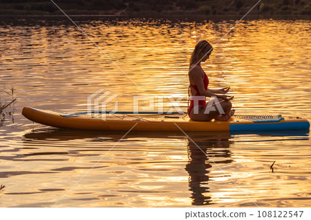 Young woman in yoga pose practicing on paddle sup surfboard during sunrise or sunset. 108122547