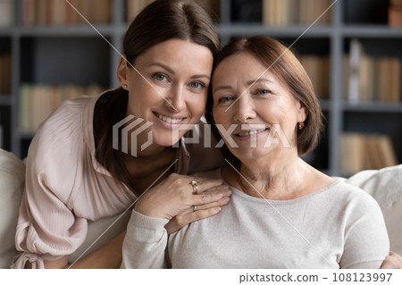 Adult daughter hugs from behind seated on sofa elderly mother Adult daughter hugs from behind seated on sofa elderly mother 108123997