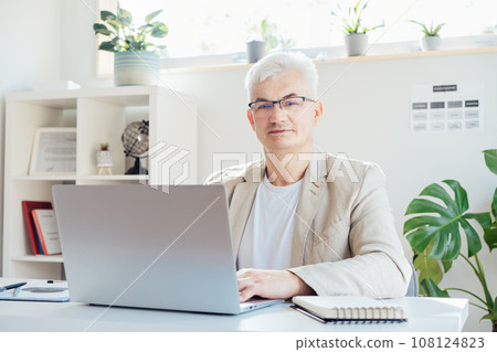 Portrait of smiling grey hair middle-aged man working on laptop while sitting at his work place in office. Confident, experienced senior male professional. Small business entrepreneur manage business 108124823