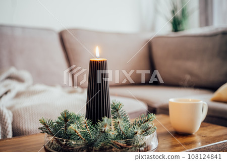Composition of flaming black christmas candle and spruce branches and cup with hot drink on coffee table in living room. Natural Christmas decorations for home. Cozy Christmas winter domestic mood. 108124841