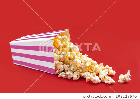Tasty cheese popcorn falling out of a pink striped carton bucket, isolated on red background Tasty cheese popcorn falling out of a pink striped carton bucket, isolated on red background 108125670
